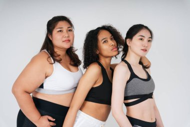 Beyaz arka planda spor sutyen giyen Asyalı ve Afrikalı kadınların grup portresi.