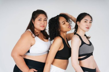 Beyaz arka planda spor sutyen giyen Asyalı ve Afrikalı kadınların grup portresi.