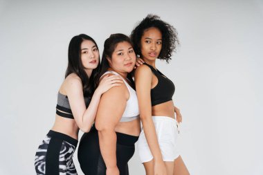 Beyaz arka planda spor sutyen giyen Asyalı ve Afrikalı kadınların grup portresi.