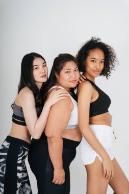 Beyaz arka planda spor sutyen giyen Asyalı ve Afrikalı kadınların grup portresi.