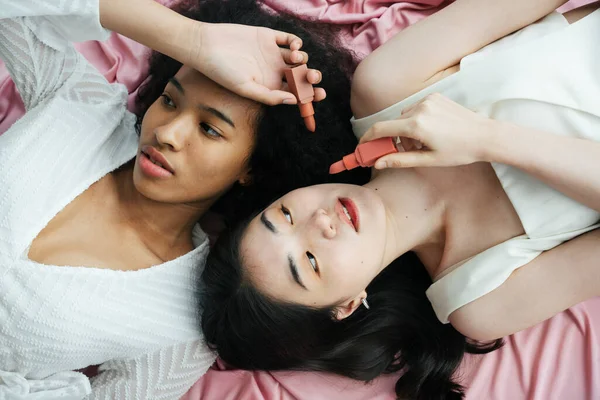 Üst Manzara - Beyaz gömlekli Asyalı ve Afrikalı kadınlar Pembe kumaş üzerinde ruj tutuyorlar.