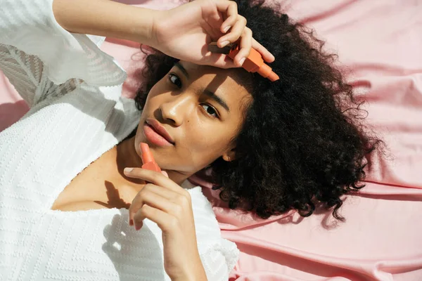 Üst Manzara - Pembe kumaş arka planda kırmızı ruj tutan Afrikalı kıvırcık saçlı kadın.