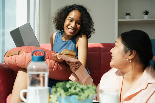African and asian women living together at home. Chubby woman share donut with friend.