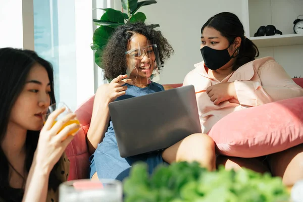 Diversity society concept. Asian and african women wear face shield and mask using laptop at home.