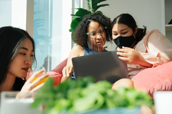 Diversity society concept. Asian and african women wear face shield and mask using laptop at home.