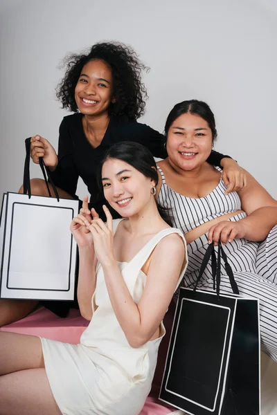 Asyalı ve Afrikalı üç farklı kadının grup portresi beyaz arka planda taklit etmek için alışveriş torbaları tutuyor..