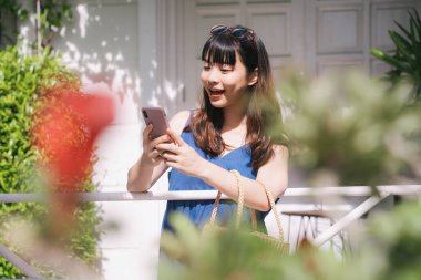 Mavi elbise giyen neşeli genç Asyalı kadın gezgin otel rezervasyonu yapıyor ya da internetten akıllı telefonlu şeyler sipariş ediyor..