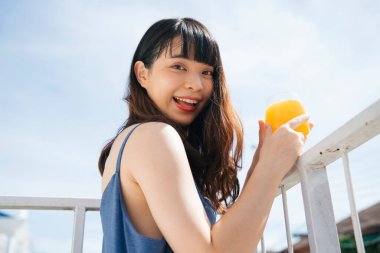 Mavi elbiseli genç Asyalı kadın gezgin balkonda sağlıklı portakal suyu içerek eğleniyor..