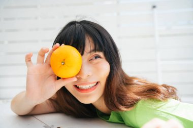 Yeşil tişörtlü genç Asyalı kadın gezgin gözlerinde portakal meyvesi tutuyor..