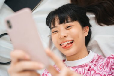 Üst Manzara - Pembe tişörtlü genç Asyalı kadın yatakta akıllı telefon kullanmaktan hoşlanıyor.