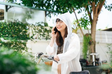 Bisikletli akıllı telefon kullanan beyaz yağmurluk giyen genç Asyalı kadın..