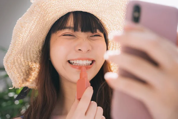 特写亚洲女旅行家戴帽子 涂口红 用智能手机照镜子的照片 — 图库照片