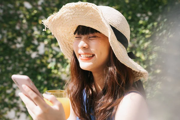 若い陽気なアジアの女性旅行者は スマートフォンを使用してわら帽子をかぶってオレンジジュースのガラスを保持 — ストック写真