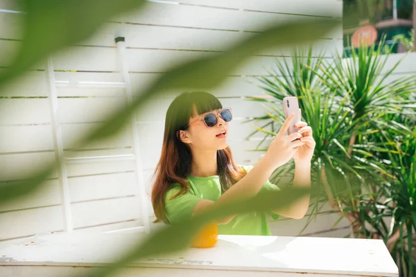 年轻的亚洲女性旅行者戴着太阳镜 坐在酒吧里喝橙汁 在院子里用智能手机 — 图库照片