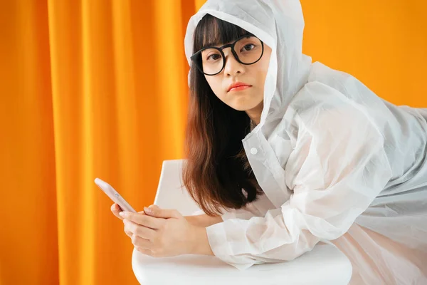 비옷을 안경을 이시아 여성이 주황색 배경에 스마트폰을 사용하는 — 스톡 사진