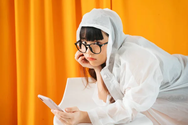 비옷을 안경을 이시아 여성이 주황색 배경에 스마트폰을 사용하는 — 스톡 사진