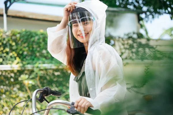 白いレインコートとフェイスシールドを身に着けている若いアジアの女性は屋外公園で自転車に — ストック写真