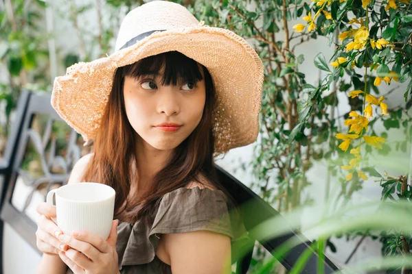 若いですアジアの女性を身に着けていますわら帽子保持カップのコーヒーで庭屋外 — ストック写真