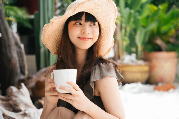 年轻的亚洲女人戴着帽子 在户外的院子里端着一杯咖啡 — 图库照片