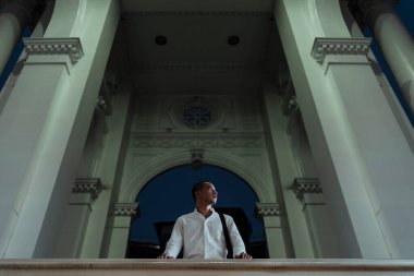 Beyaz tişörtlü dazlak adamın beyaz mimarinin önünde durduğu düşük açılı bir kare..