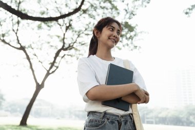 Kitabı tutan bir kız, üniversitenin bahçesinde duruyor ve gülümsüyor..