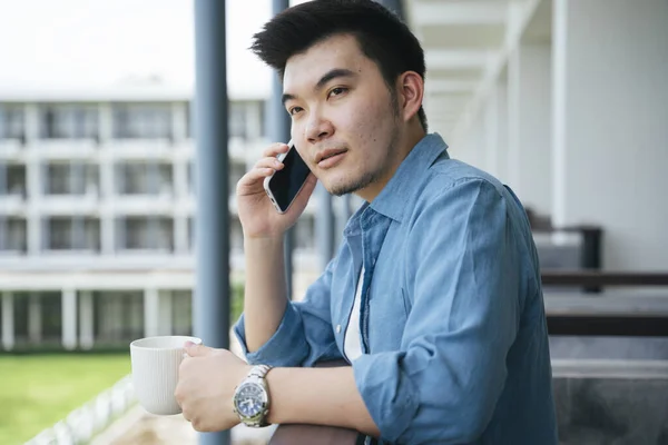 Asian business man in blue collar shirt talking on the phone at hotel balcony.