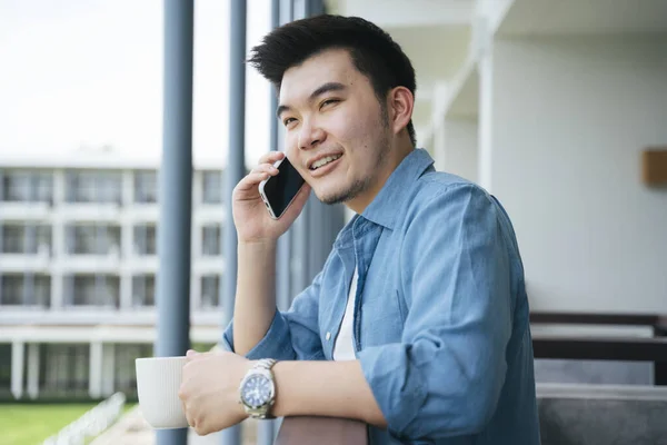 Asian business man in blue collar shirt talking on the phone at hotel balcony.