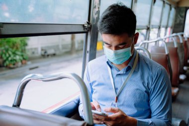 Asyalı iş adamı otobüste akıllı telefon kullanarak maske takar..
