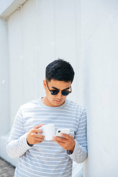 Portrait of stylish asian adult man wear sunglasses using smartphone and hold a cup of coffee.