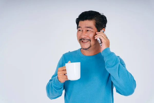 老年男子穿着蓝色长袖衬衫 手持白咖啡杯在电话中交谈的画像 — 图库照片