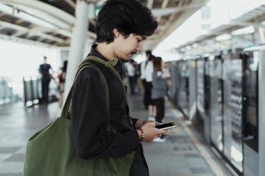 Siyah saçlı adam tren istasyonunda sakince duruyor ve tren beklerken cep telefonunu kullanıyor..