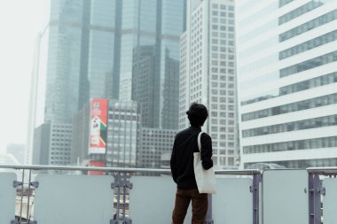 Beyaz çantalı siyah saçlı adam gökyüzü tren istasyonunun terasında duruyor. Şehirdeki yüksek camlı binaya bak..