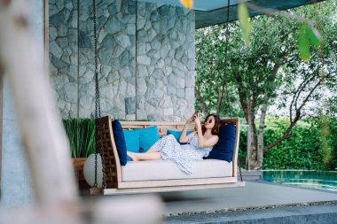 Güzel Asyalı gezgin telefon kullanıyor. Havuz kenarındaki salıncakta dinleniyor.