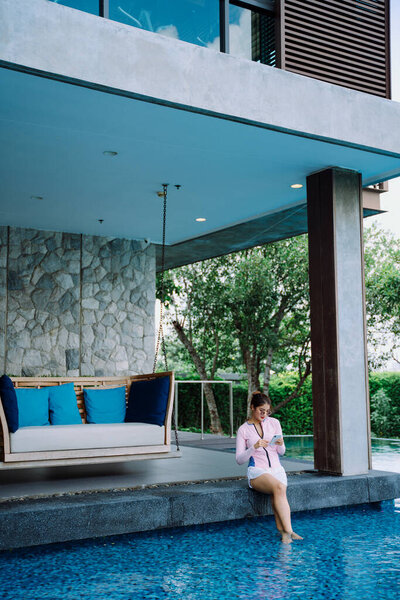 Asian woman traveler wear swim suit using phone at poolside in resort and hotel.