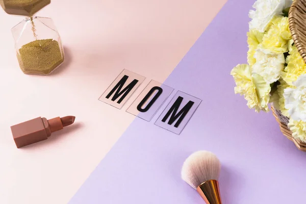 Mom word with lipstick and brush and sand glass on pastel background.