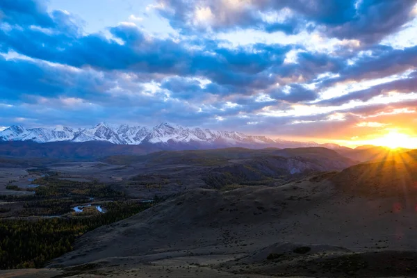 山の風景。太陽は山の後ろに行き、地平線の近くの丘と白い峰を照らします。アルタイ共和国. — ストック写真