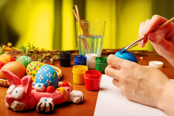 The artist paints Easter religious items. Chicken egg, prepared for coloring, is in the basket. Branches of willow and lilac with blooming green leaves. Paint in a jar with brushes and a glass of water. Hands close up