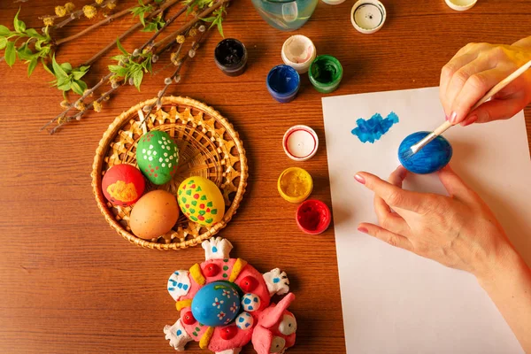 The artist paints Easter religious items. Chicken egg, prepared for coloring, is in the basket. Branches of willow and lilac with blooming green leaves. Paint in a jar with brushes and a glass of water. Hands close up