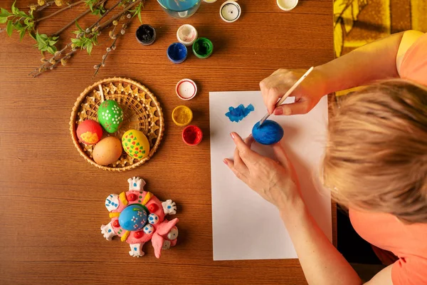 The artist in glasses performs design work. Paints eggs with paints. Spring, warm mood. On the table are the branches of willow and lilac with green flowering leaves. Multicolored Easter still life