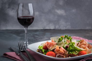 Prosciutto ve parmesan ile salata. Çatal bıçak, bıçak, çatal ve gri arka plan üzerinde pembe peçete. Bir kadeh kırmızı şarap