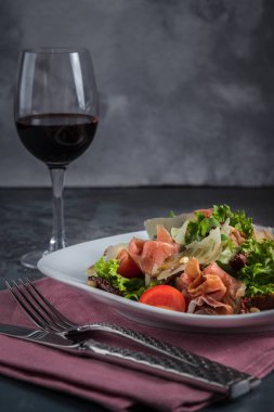 Prosciutto ve parmesan ile salata. Çatal bıçak, bıçak, çatal ve gri arka plan üzerinde pembe peçete. Bir kadeh kırmızı şarap