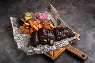 Minsk, Beyaz Rusya - Ağustos, 2019 Barbekü Domuz kaburga pişmiş patates ve salatalık turşusu ile ayarlanmış duman bir restoranda gri bir beton masa üzerinde yalan ahşap arka plan tahtası üzerinde gazete ile hizmet