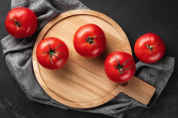 many red and fresh raw tomatoes in the middle of a round wooden serving plate with tablecloth. dark black and gray textured background, top view