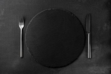 Çatal ve bıçak manzaralı yuvarlak siyah granit taş tahta. Metnin için kopyalama alanı olan siyah desenli çimento arkaplan ve şablon