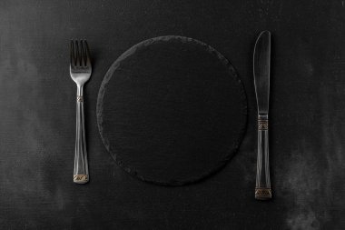 Yuvarlak, siyah granit taş tahta, parlak çatal ve bıçak üst görüşlü. Metnin için kopyalama alanı olan siyah desenli çimento arkaplan ve şablon
