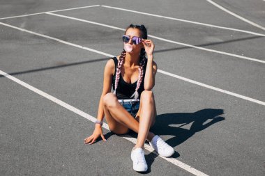 Genç güzel kadın genç kız dinlemek müzik açık kulaklık kulaklık akıllı telefonda oturan çim üzerinde otururken spor stadyumunun üzerinde oturmuş pembe saç cam krem yiyecek