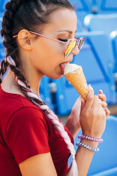 Young beautiful woman teen girl listen music outdoor earphones headphone on smartphone lying on the grass sitting on sport stadium attractive braided pink hair braids bubble gum summer hot pink glasses eating ice cream