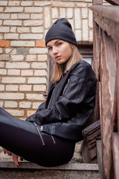 стильная красивая девушка блондинка позирует прогулки по городу в черной шляпе, черная кожаная куртка и брюки, высокие сапоги. Портрет. сидя на ступеньках
.