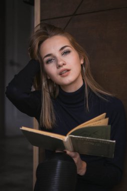 Bir kafede oturan, kitap okuyan, kahve içen genç güzel kız. Kameraya poz vermek. Siyah bir kazak ve siyah şapka giymiş. Sarı saçlı, gülümseyen, tatlım. Sonbahar eğlencesi