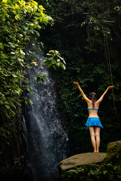 Взволнованная белая женщина поднимает руки перед водопадом. Вид сзади. Возле Тегенунганского водопада. Убуд, Бали, Индонезия — стоковое фото